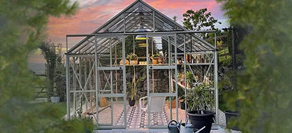 A glass Rhino Greenhouse showcases various plants on shelves, surrounded by a garden setting with trees and potted plants, under a colorful sunset sky.