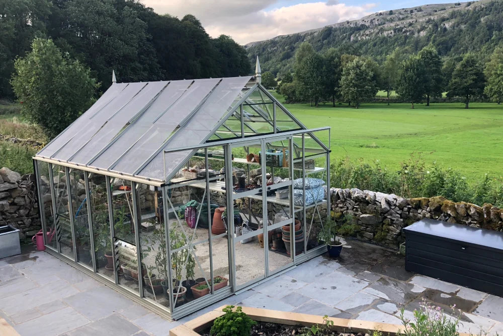 A glass Rhino Greenhouse stands filled with potted plants and gardening tools, surrounded by stone walls and lush green fields, set against a backdrop of trees and hills.