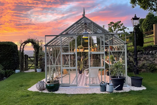 A glass Rhino Greenhouse stands, filled with plants and a white chair, on a neatly gravelled area surrounded by lush grass and a stone wall, under a vibrant sunset sky.