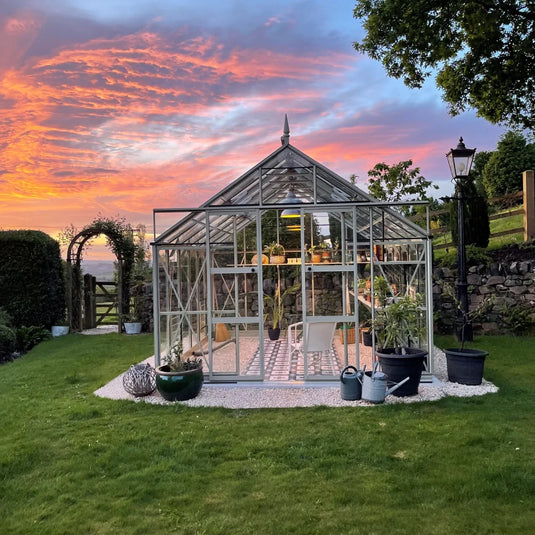 A glass Rhino Greenhouse stands illuminated, displaying potted plants inside. It's situated in a grassy garden, surrounded by a stone wall, trees, and lamp post under a vibrant sunset sky.