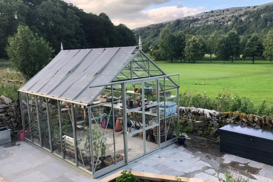 A glass Rhino Greenhouse stands with plants and tools inside, surrounded by stone walls. It's set in a scenic landscape with lush green fields and trees, and distant hills.