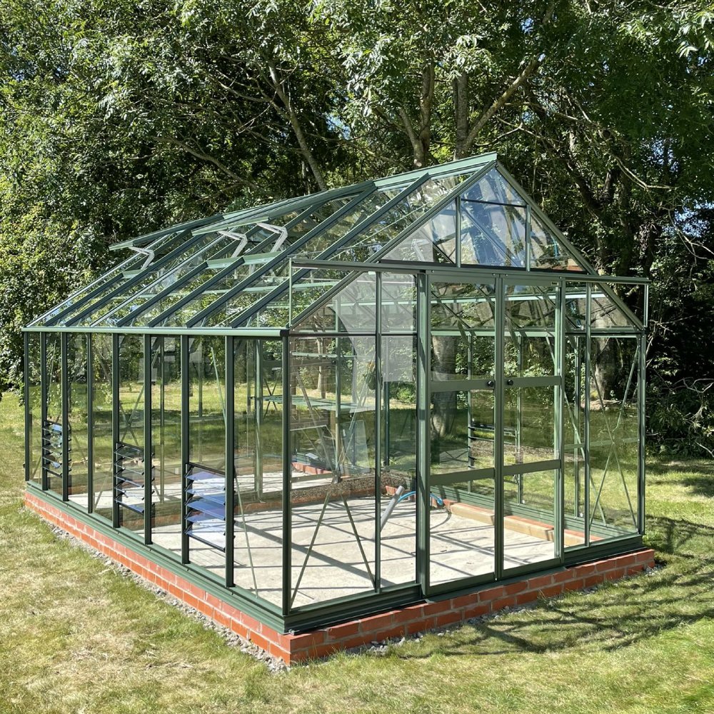 A green metal-framed Rhino Greenhouse stands on a brick foundation, surrounded by lush trees and grass. Sunlight illuminates its glass panels, creating a bright, inviting atmosphere.
