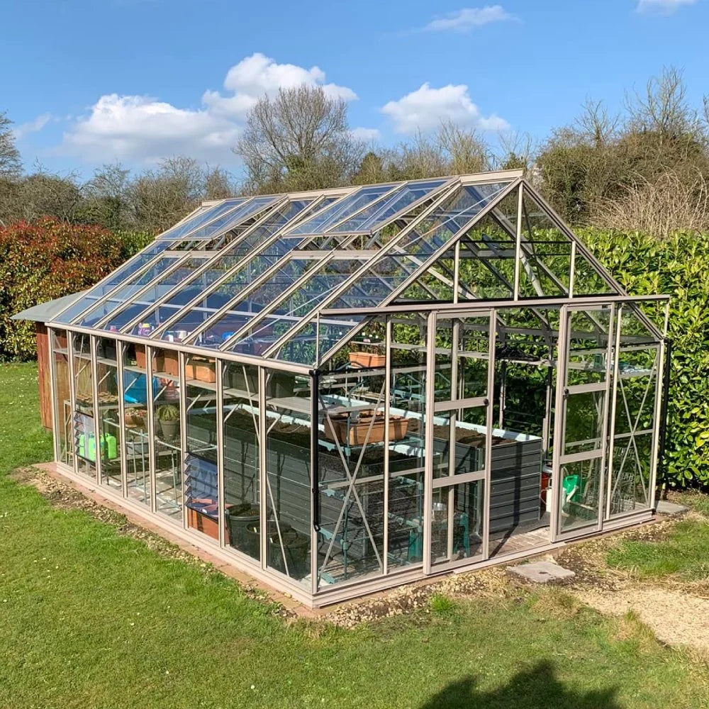 A large glass Rhino Greenhouse sits on a grassy lawn, surrounded by trees and hedges, with gardening tools and plants visible inside under a clear blue sky.