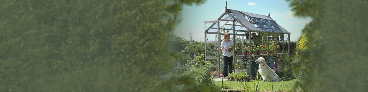 A Rhino Greenhouse holds potted plants, with a person tending them and a dog sitting nearby. The setting is a lush garden with dense green foliage.