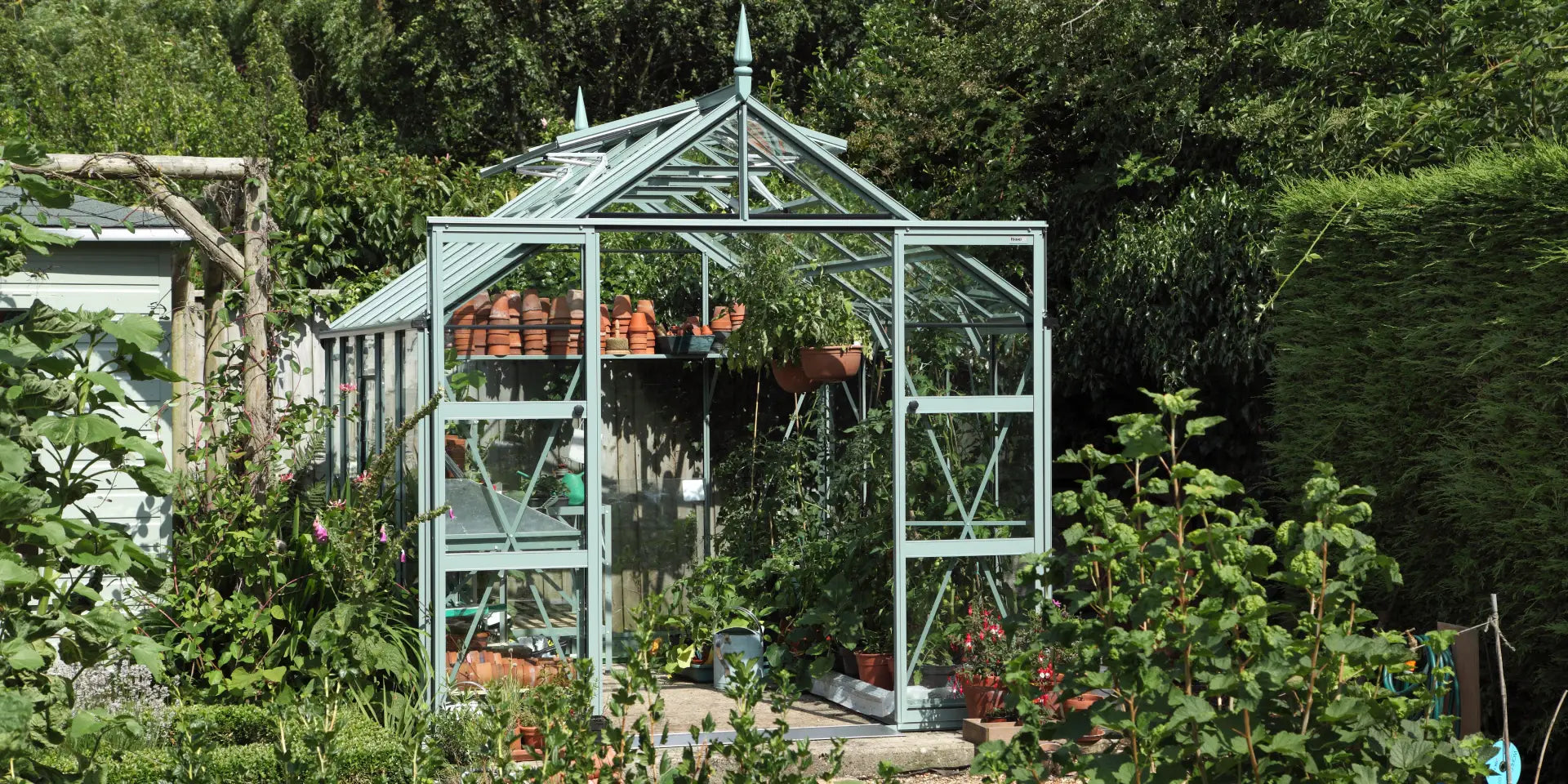 Rhino Greenhouse filled with plants and terracotta pots, surrounded by lush greenery in a garden setting. The structure features glass panels and a pointed roof.