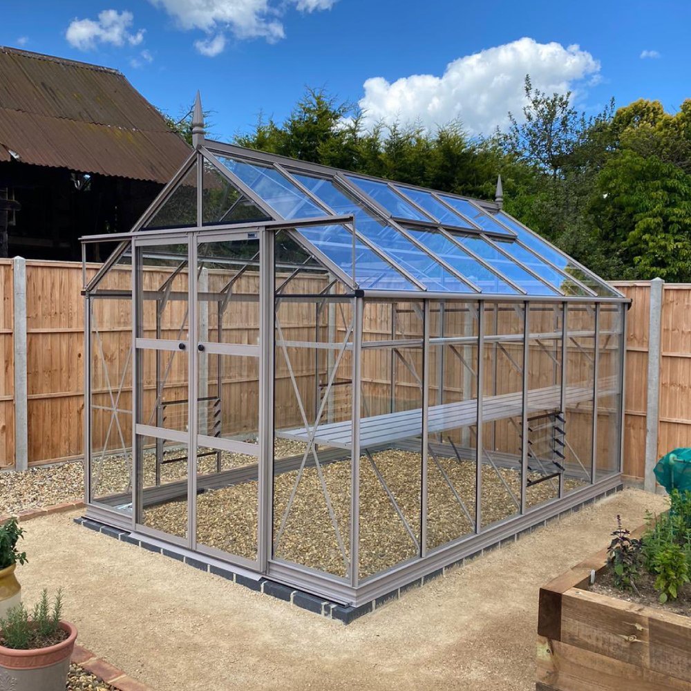 A glass Rhino Greenhouse stands on gravel, surrounded by a wooden fence and garden beds, under a blue sky with clouds.