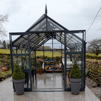 A glass Rhino Greenhouse stands open, revealing potted plants and gardening tools on shelves. It is situated outdoors, surrounded by stone walls and a rural landscape under a cloudy sky.
