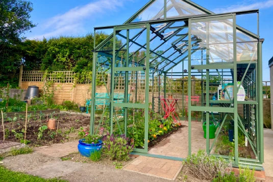 A green Rhino Greenhouse stands open, housing various plants and gardening tools, amidst a vibrant garden with a blue planter and lush foliage surrounding the area.