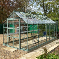 A glass Rhino Greenhouse stands on a tiled base, surrounded by gravel and garden foliage, with large windows reflecting sunlight, and trees visible in the background.