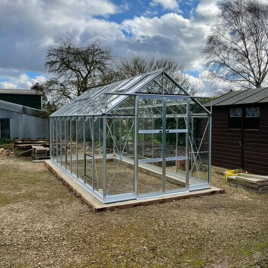 A large, glass-paneled Rhino Greenhouse stands on a concrete base, surrounded by trees and adjacent to a wooden shed under a partly cloudy sky.