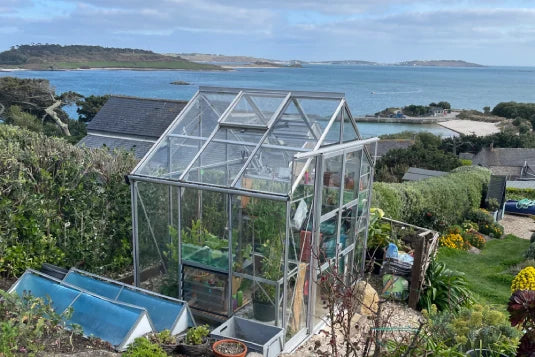 A glass Rhino Greenhouse stands on a lush hillside, housing plants inside. The setting overlooks a scenic ocean view, with houses and gardens visible in the background.