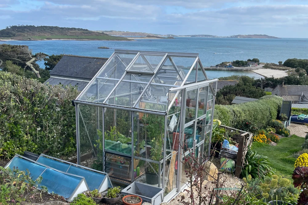 A Rhino Greenhouse with glass panels stands in a lush garden filled with plants, overlooking a scenic coastline with blue water and distant islands under a partly cloudy sky.
