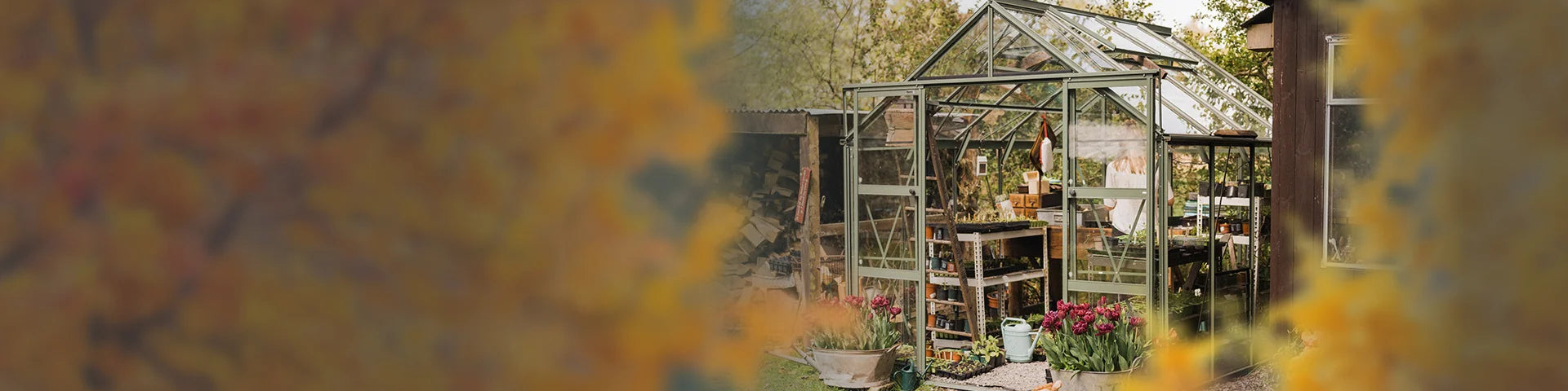 Glass Rhino Greenhouse with open doors, housing gardening tools and potted plants. It is situated in a garden, surrounded by trees and vibrant flowers in the foreground.