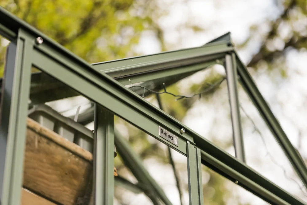 Green metal Rhino Greenhouse frame with a Rhino label, partially open roof vent. Background includes blurred green foliage and sky, suggesting an outdoor garden setting.