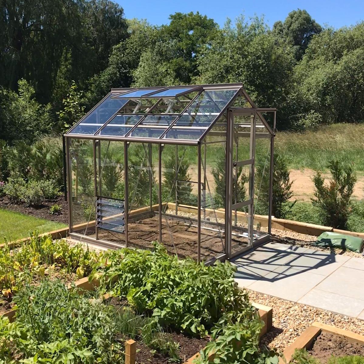 A glass Rhino Greenhouse stands on a paved area, surrounded by lush garden beds and trees under a clear blue sky.