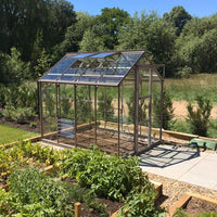 A glass Rhino Greenhouse stands on a paved area, surrounded by lush garden beds and trees under a clear blue sky.
