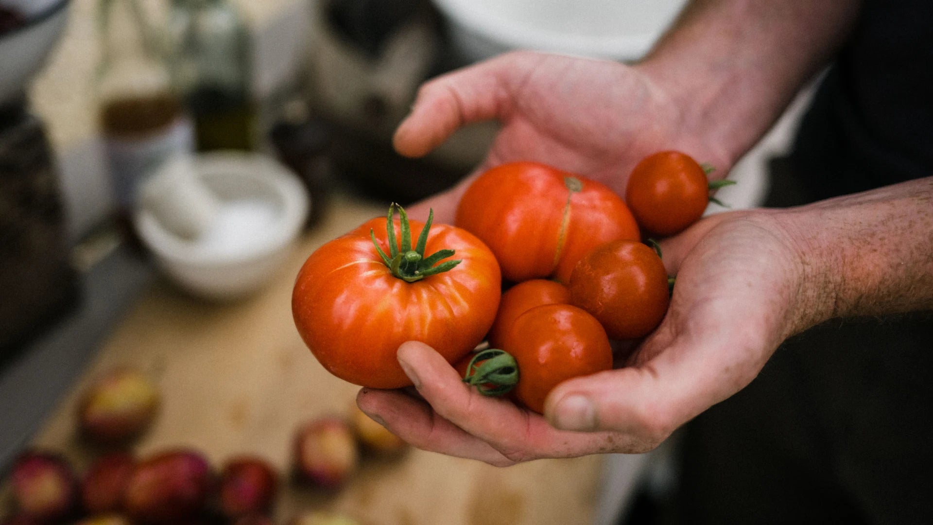 Hands holding ripe tomatoes, surrounded by a kitchen setting with blurred bowls and jars in the background.