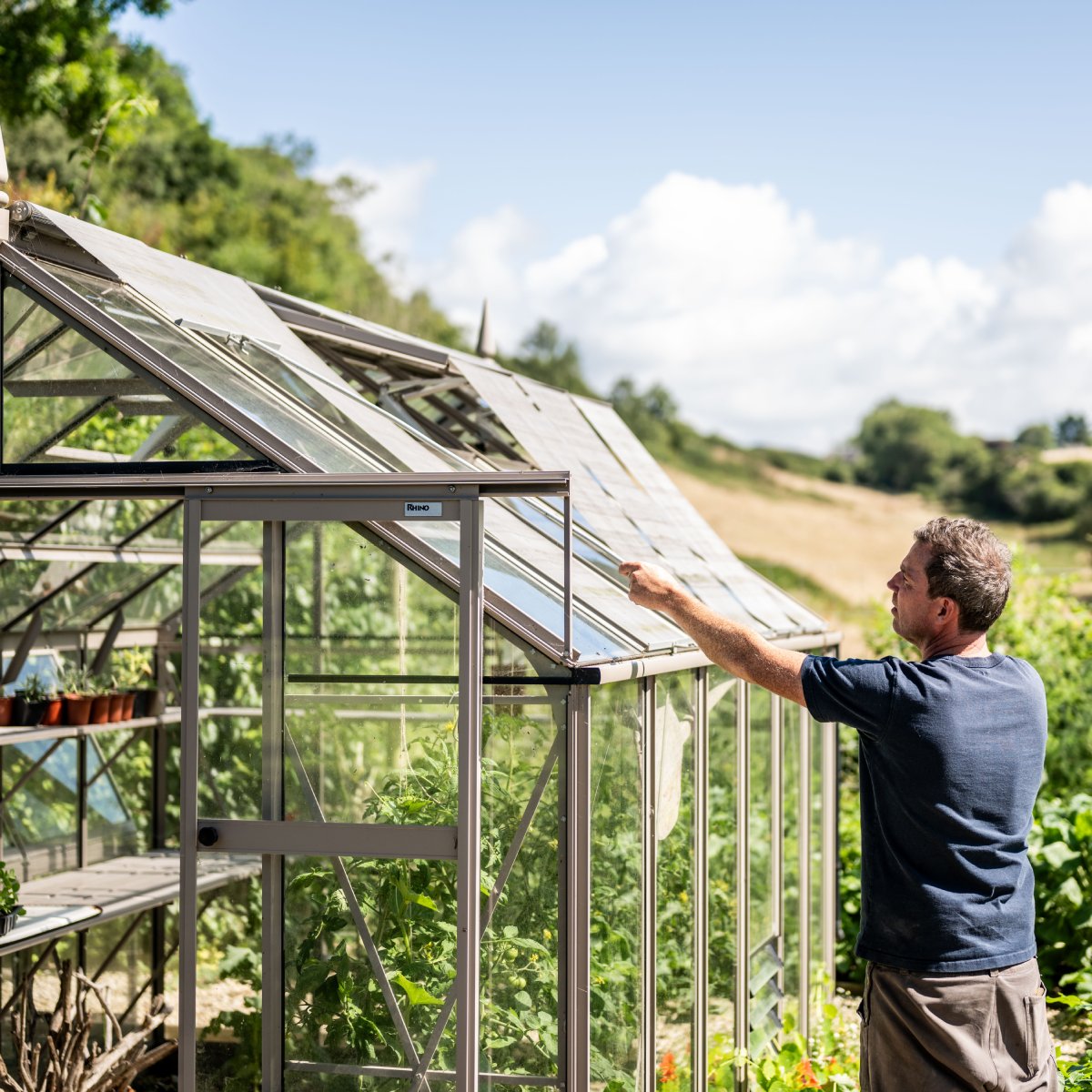 A man adjusts a window on a glass Rhino Greenhouse labeled Rhino, surrounded by green plants and countryside under a blue sky.