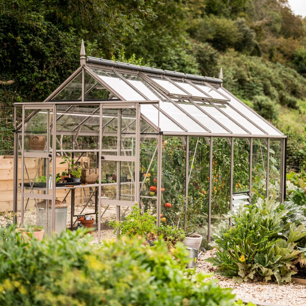 A glass Rhino Greenhouse stands with plants and gardening tools inside, situated in a lush garden setting surrounded by trees and various greenery.