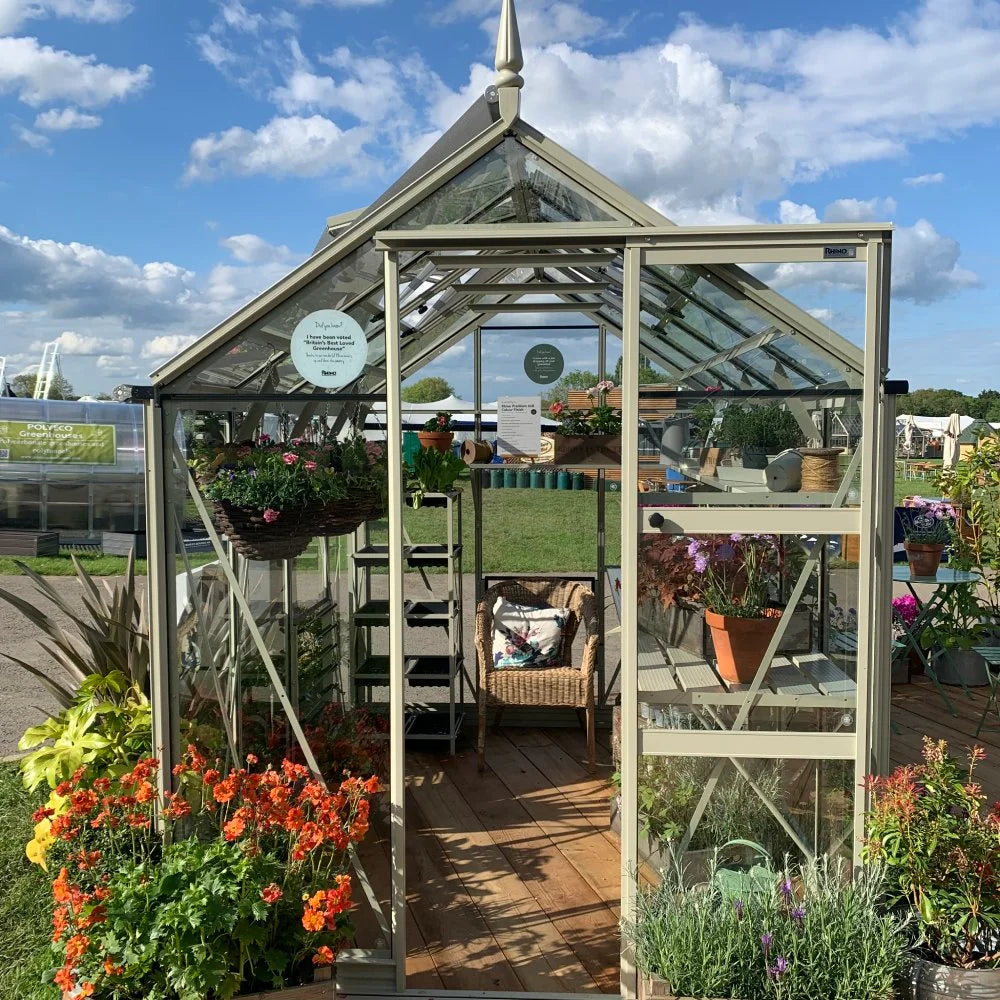 A glass Rhino Greenhouse with an open door displays vibrant potted plants and hanging baskets inside. A wicker chair sits on the wooden floor, surrounded by a grassy outdoor setting. Signs inside the Rhino Greenhouse read, Designed and manufactured in Yorkshire. A Rhino Rhino Greenhouse is a Rhino Greenhouse for life, and Buy now.