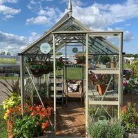 A glass Rhino Greenhouse with an open door displays vibrant potted plants and hanging baskets inside. A wicker chair sits on the wooden floor, surrounded by a grassy outdoor setting. Signs inside the Rhino Greenhouse read, Designed and manufactured in Yorkshire. A Rhino Rhino Greenhouse is a Rhino Greenhouse for life, and Buy now.
