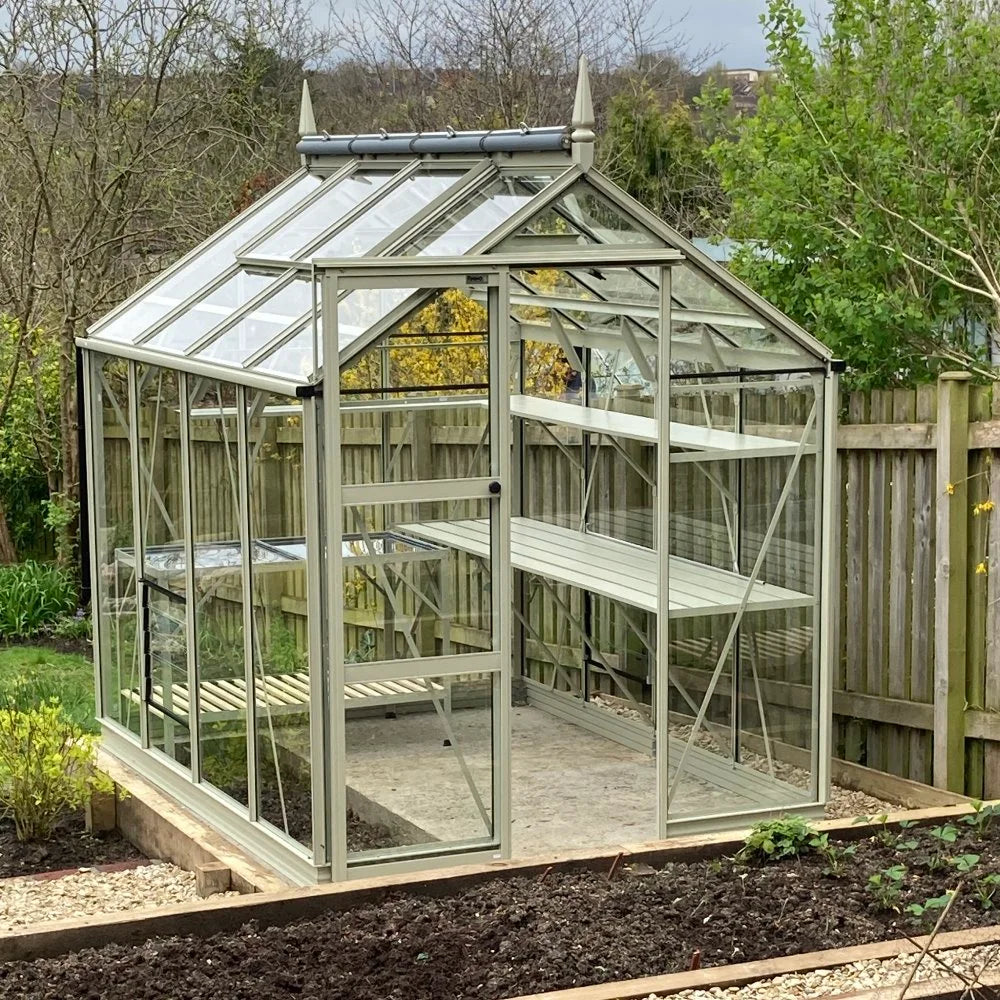 A glass Rhino Greenhouse stands on a concrete base with internal shelves, surrounded by a wooden fence, leafless trees, and a garden plot filled with soil.