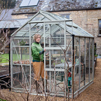 A person is standing inside a glass Rhino Greenhouse, sliding its door open. The Rhino Greenhouse is situated in a garden with a stone building in the background. The text Rhino is visible.