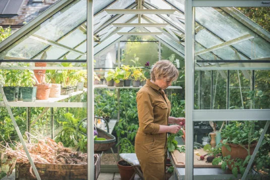 A person tends to plants in a glass Rhino Greenhouse filled with various potted greenery and gardening tools, under natural light filtering through transparent panels.