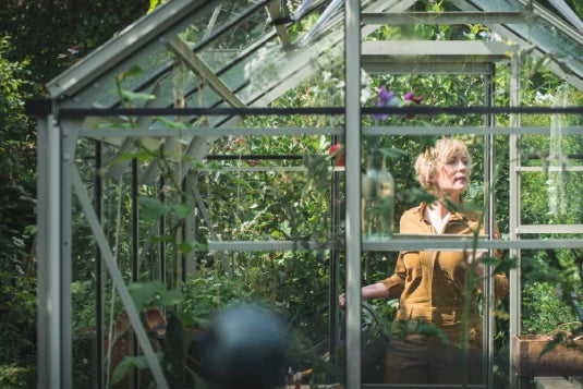 A person stands inside a glass Rhino Greenhouse, tending plants, surrounded by lush greenery. Sunlight filters through the glass, highlighting the vibrant vegetation and intricate Rhino Greenhouse structure.
