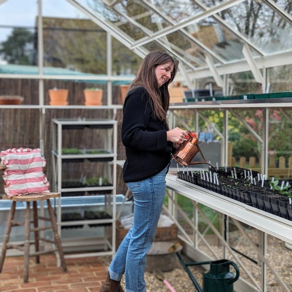 A woman waters plants with a copper watering can inside a Rhino Greenhouse. Surrounding shelves hold potted plants and gardening tools, creating a cozy gardening environment.