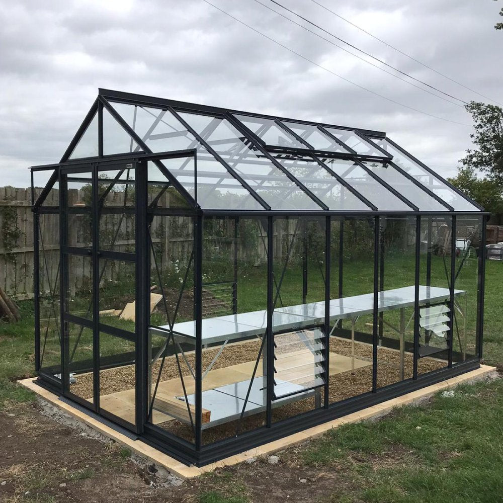 A glass and metal Rhino Greenhouse stands on a wooden base, with vented windows and internal shelves, situated in a grassy area enclosed by a wooden fence under a cloudy sky.