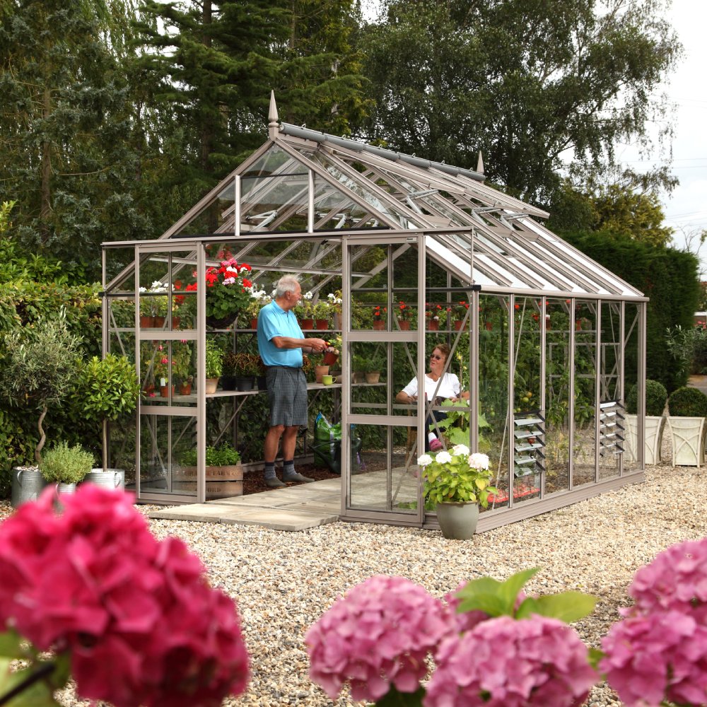 A Rhino Greenhouse shelters colorful plants inside, where two people interact. It stands on gravel, surrounded by greenery and pink blossoms, under a canopy of large trees.