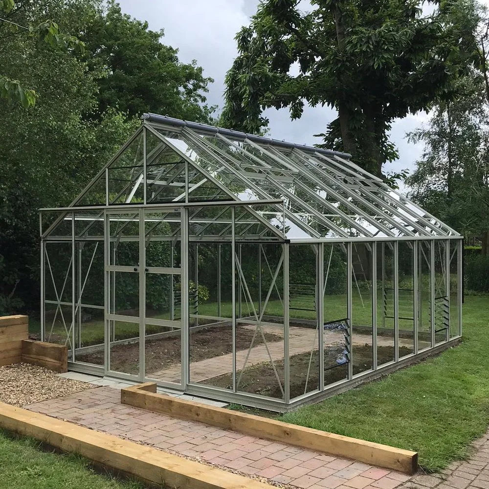 A glass Rhino Greenhouse stands on a paved path surrounded by lush greenery and trees, with a well-manicured lawn extending around the structure.