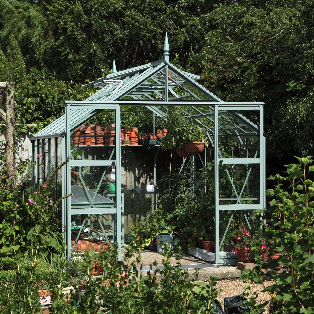 A glass Rhino Greenhouse with open doors, filled with potted plants and gardening tools, is set within a lush garden surrounded by dense greenery.