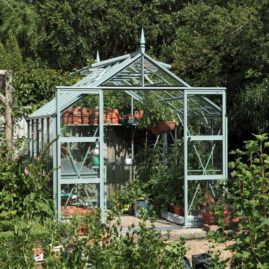 A glass Rhino Greenhouse with open doors, filled with potted plants and gardening tools, is set within a lush garden surrounded by dense greenery.