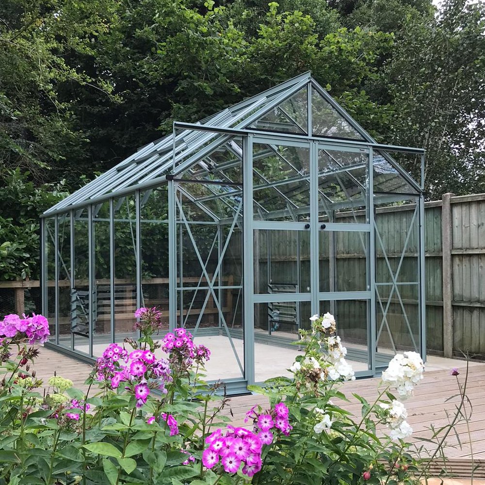 A glass Rhino Greenhouse stands on a wooden deck, surrounded by colorful pink and white flowers. Tall green trees and a wooden fence provide a natural backdrop.