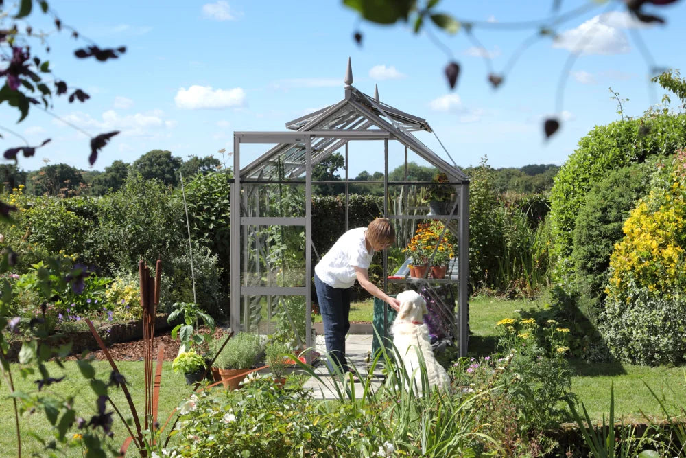 A person is bending to pet a dog in front of a glass Rhino Greenhouse surrounded by lush, colorful garden plants on a sunny day.