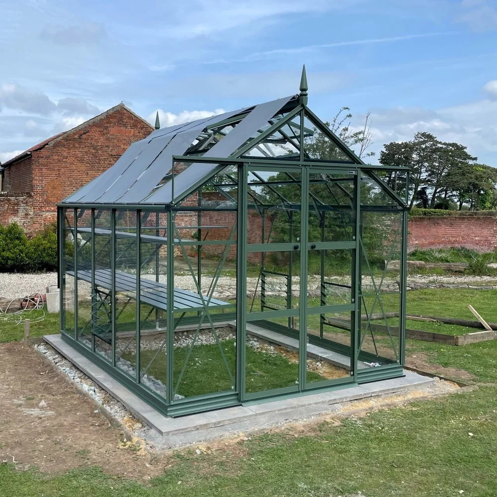 A green metal-framed Rhino Greenhouse sits on a concrete base, with sliding doors open. It's in a garden with a brick wall and trees in the background, under a clear sky.