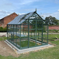 A green metal-framed Rhino Greenhouse sits on a concrete base, with sliding doors open. It's in a garden with a brick wall and trees in the background, under a clear sky.