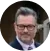 A person with glasses and facial hair stands outdoors, wearing a formal outfit. The background features a building and greenery in a blurred setting.