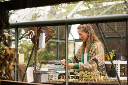 Woman tending plants at table inside glass Rhino Greenhouse, surrounded by gardening tools and potted seedlings.