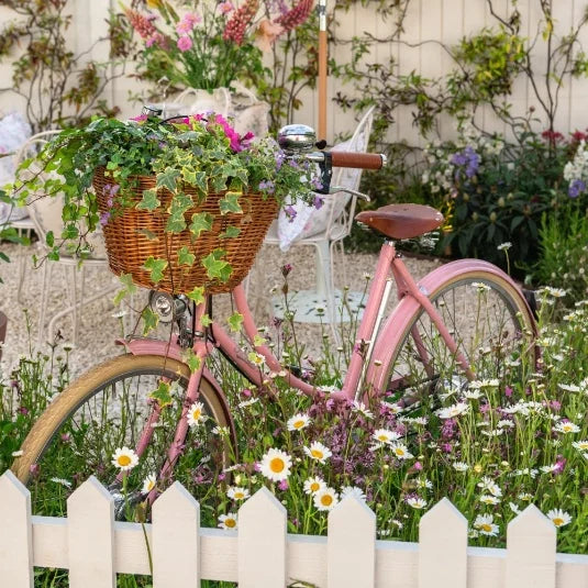 A vintage pink bicycle with a wicker basket filled with flowers and ivy stands amid wildflowers, surrounded by a white picket fence in a charming garden setting.