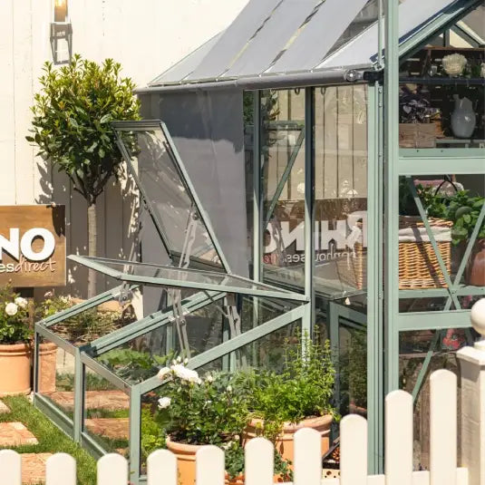 A glass Rhino Greenhouse displays potted plants and gardening tools, set against a white fence with the text RHINO Rhino Greenhousesdirect in the foreground. A neatly trimmed lawn surrounds the structure.