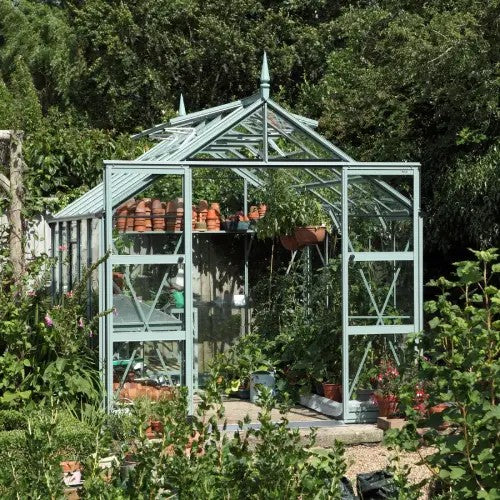 A glass Rhino Greenhouse stands amidst lush greenery, filled with potted plants and gardening tools. Sunlight filters through the structure, illuminating the thriving vegetation within a tranquil garden setting.