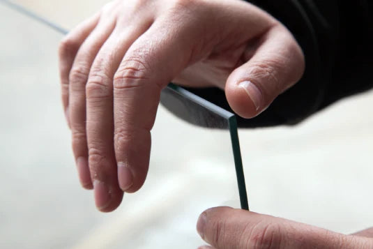 Hands hold a thin, rectangular piece of glass vertically, displaying its clarity and smooth edges against a soft, blurred background.