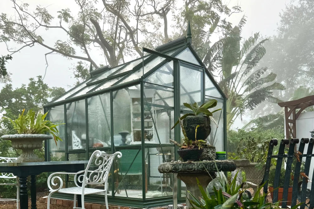 A glass Rhino Greenhouse stands surrounded by lush foliage. Inside, gardening tools and pots are visible. Outside, a wrought iron table and chairs sit among potted plants, creating a tranquil garden setting.