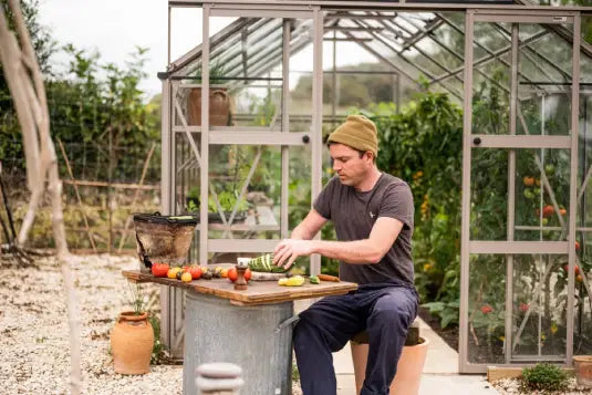 A person prepares vegetables on a table outside a Rhino Greenhouse. The context includes gardening accessories and plants, suggesting an active gardening or food preparation scene.