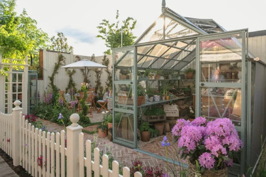 A Rhino Greenhouse with open doors displays potted plants inside. It is surrounded by a white picket fence and colorful flowers, with a nearby patio featuring a table, chairs, and umbrella.