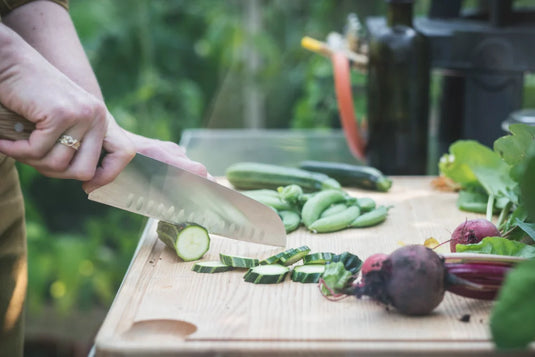 A hand slices a cucumber on a wooden cutting board surrounded by fresh vegetables like snap peas and beets, set in a garden-like environment.