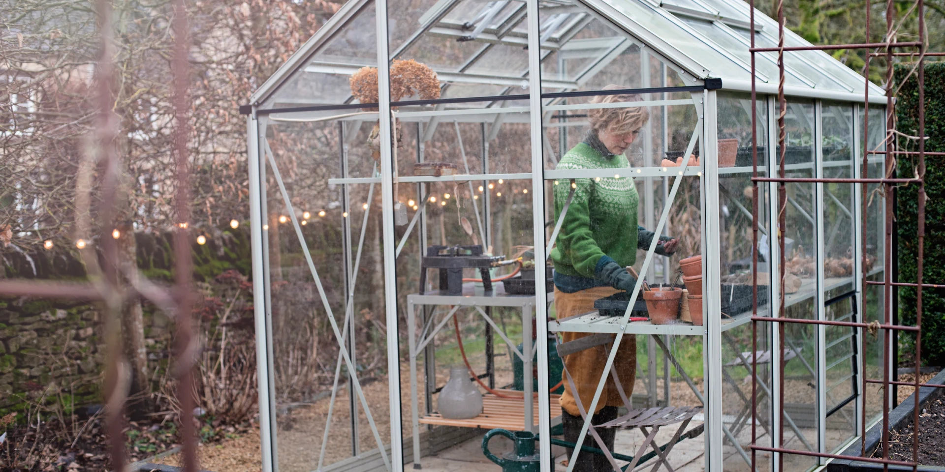 A person is potting plants inside a glass Rhino Greenhouse, surrounded by shelves with gardening tools and pots. The Rhino Greenhouse is set in a garden with trees and string lights.